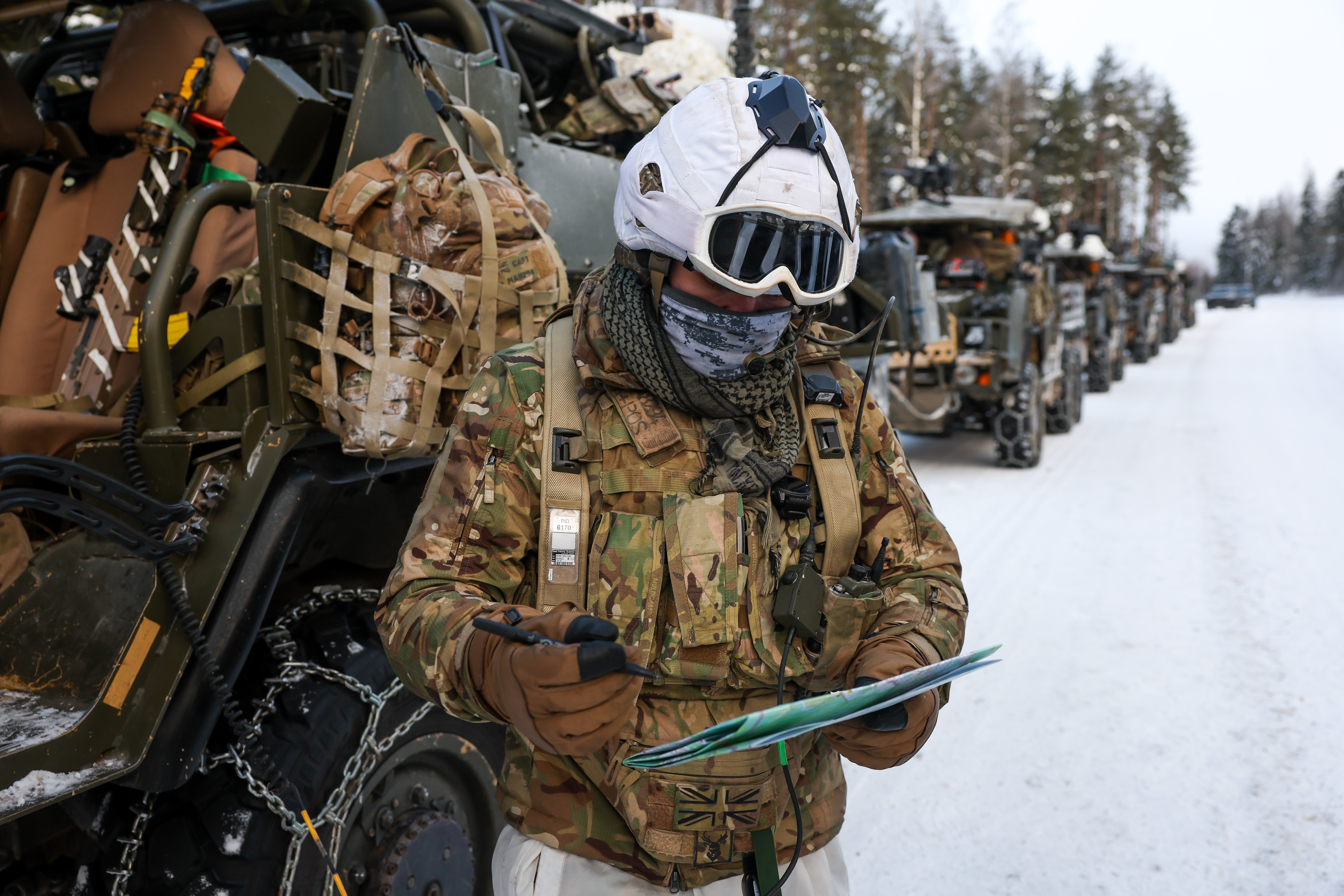 Soldier in winter camouflage gear and helmet reviews a map beside military vehicles on a snowy road.