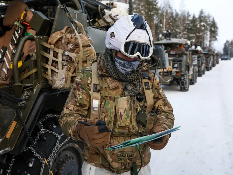 Soldier in winter camouflage gear and helmet reviews a map beside military vehicles on a snowy road.