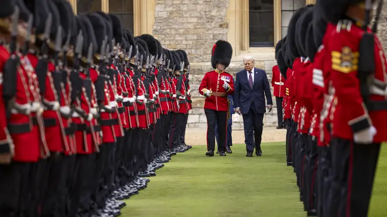 Two men walk between rows of British guards in red uniforms and bearskin hats standing to attention.