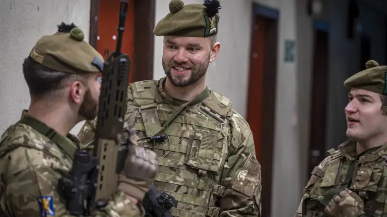 A soldier in uniform smiles and converses with another soldier who is holding a weapon.