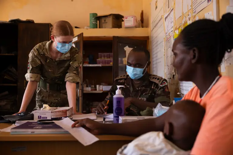 A British and Kenyan soldier are pictured with masks behind a desk, a mother looks on with her child. 