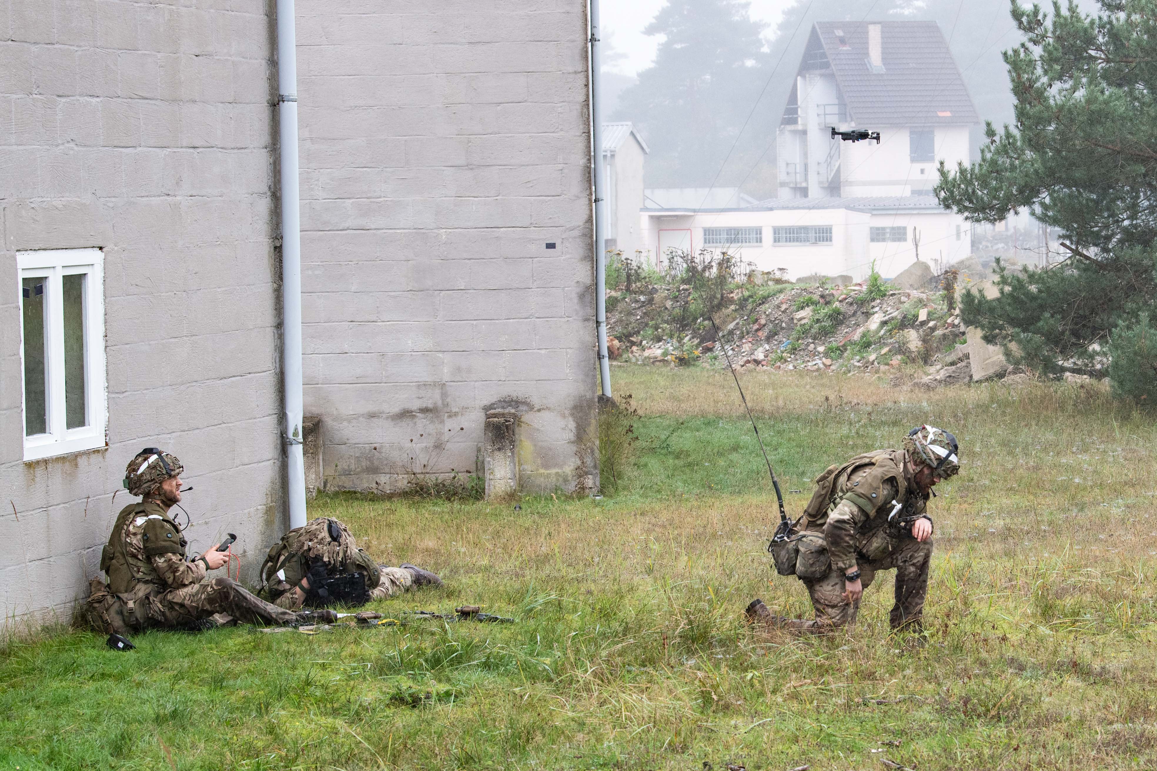 Two soldiers in camouflaged uniforms, one sitting against a building with a remote, and another kneeling, observe a hovering drone in a misty field.