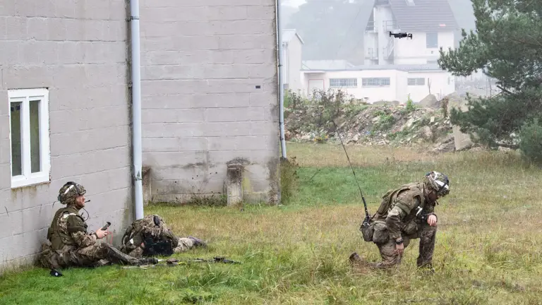 Two soldiers in camouflaged uniforms, one sitting against a building with a remote, and another kneeling, observe a hovering drone in a misty field.