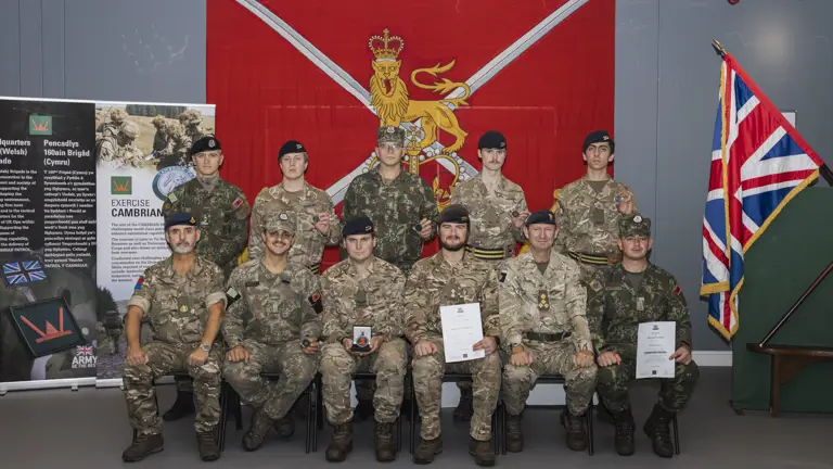 Group of military personnel in uniform posing indoors with a red flag behind them.