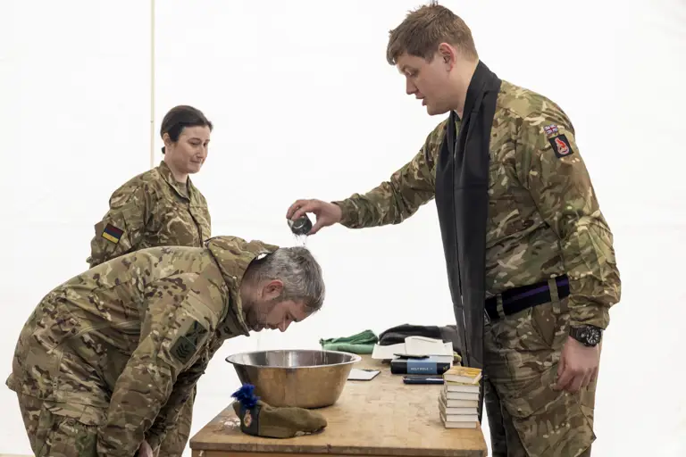 A man in uniform has water poured over his head by a Padre in Uniform during a Baptism.
