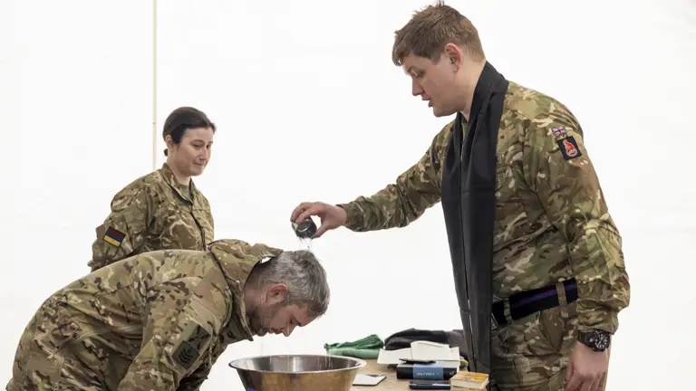 A man in uniform has water poured over his head by a Padre in Uniform during a Baptism.