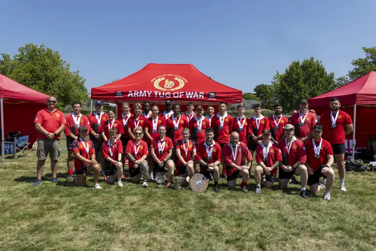 A group photo of a tug of war team, dressed in red shirts and wearing medals, posed in front of a red tent that reads 