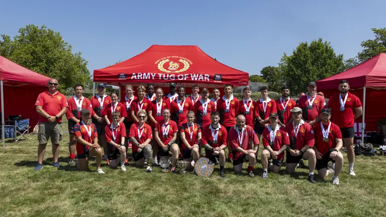 A group photo of a tug of war team, dressed in red shirts and wearing medals, posed in front of a red tent that reads