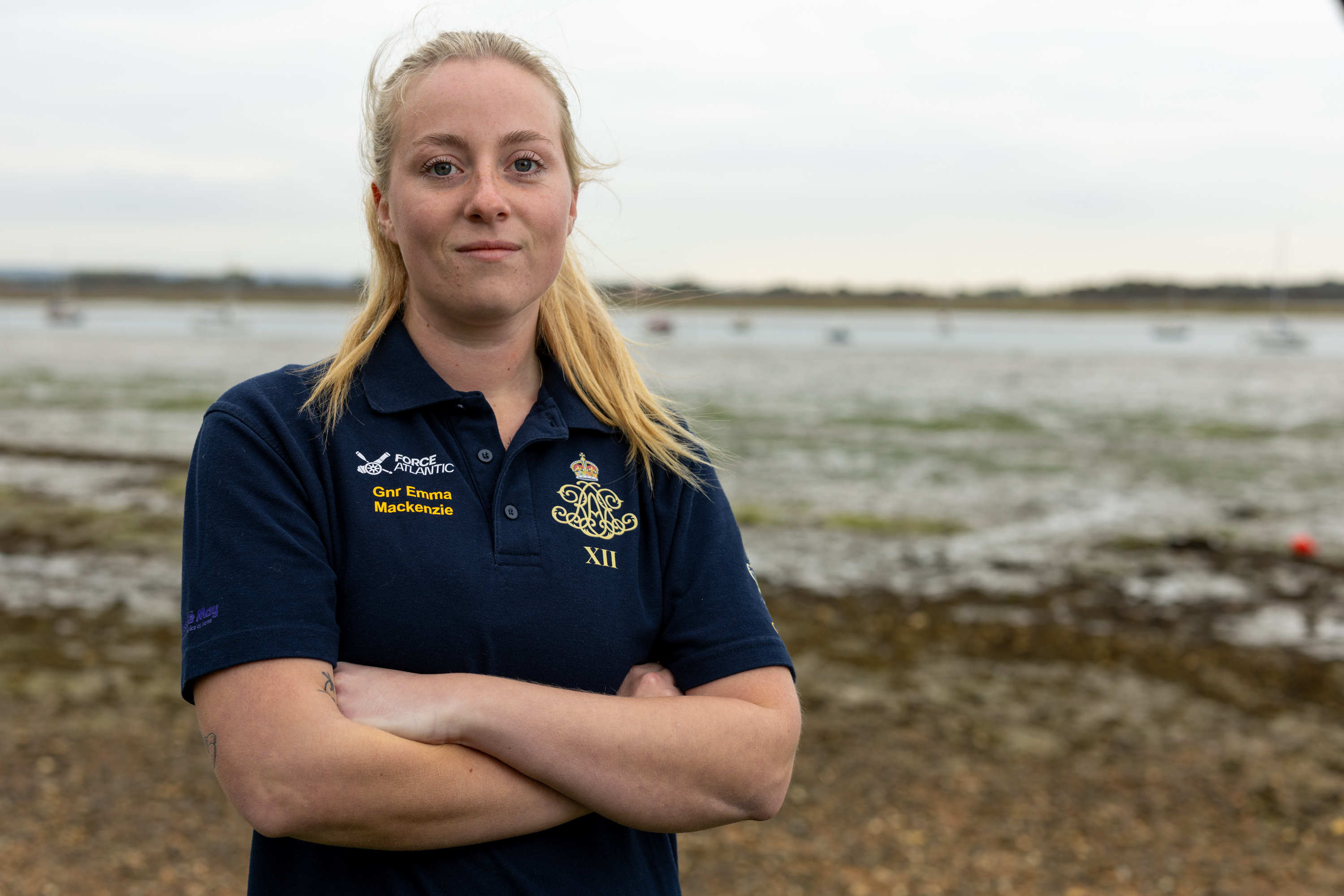 A female Army soldier, wearing dark blue shirt with an Army cap badge on it, is standing on a shoreline with an estuary in the background.