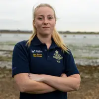A female Army soldier, wearing dark blue shirt with an Army cap badge on it, is standing on a shoreline with an estuary in the background.