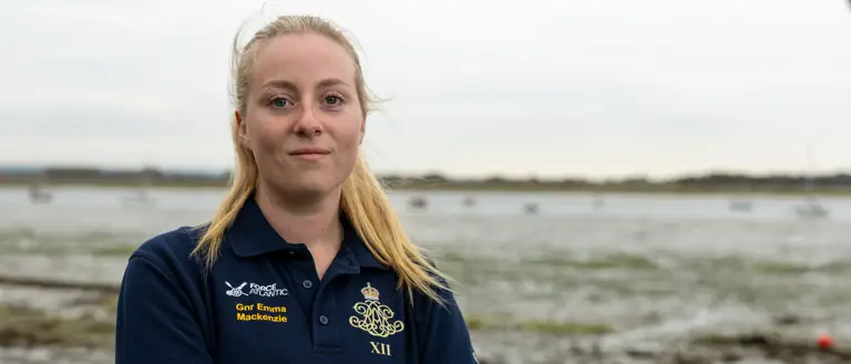 A female Army soldier, wearing dark blue shirt with an Army cap badge on it, is standing on a shoreline with an estuary in the background.