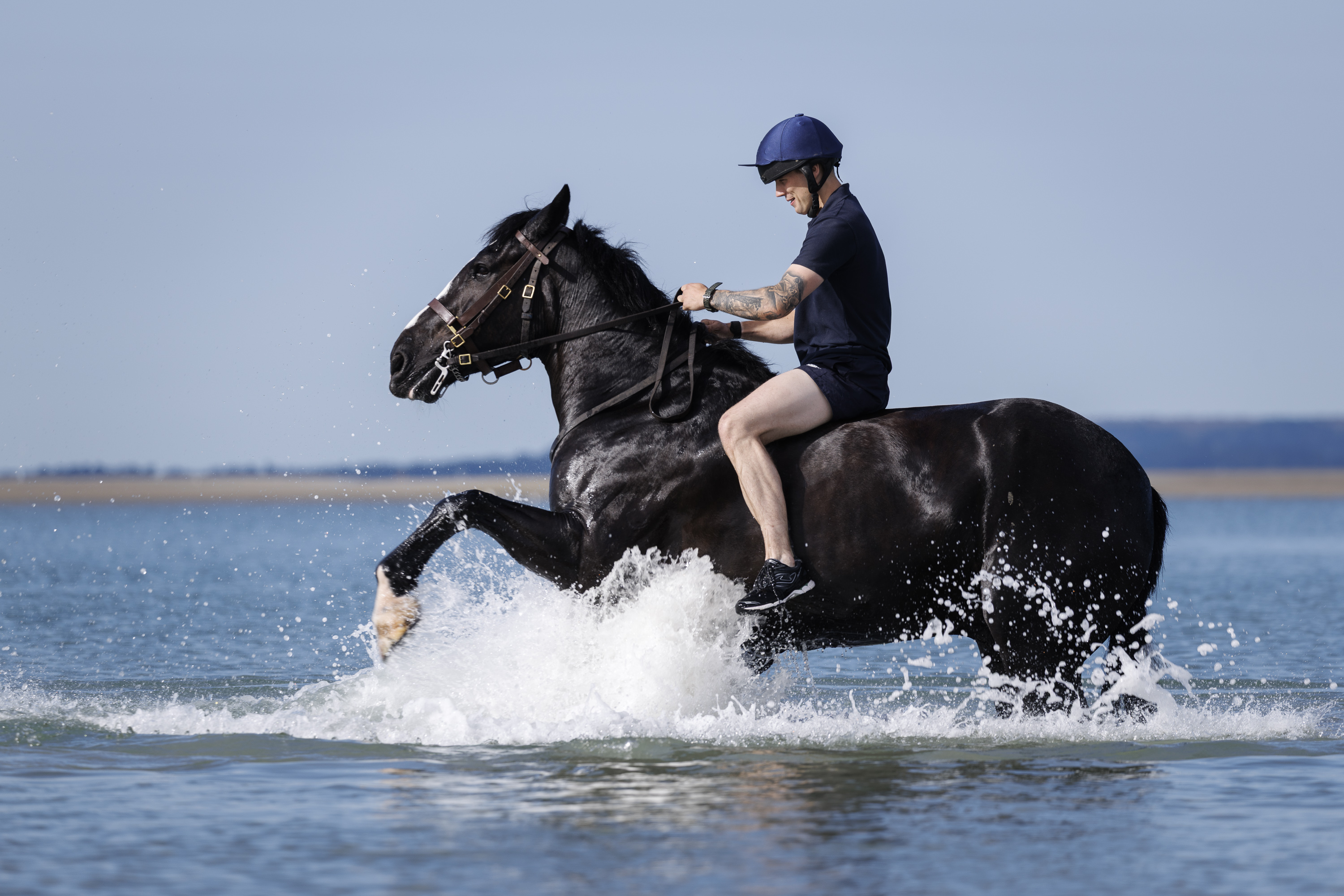 A person rides a black horse through shallow ocean water, creating splashes. The rider wears a helmet and is dressed in dark clothing. The background features a clear blue sky and distant shoreline.