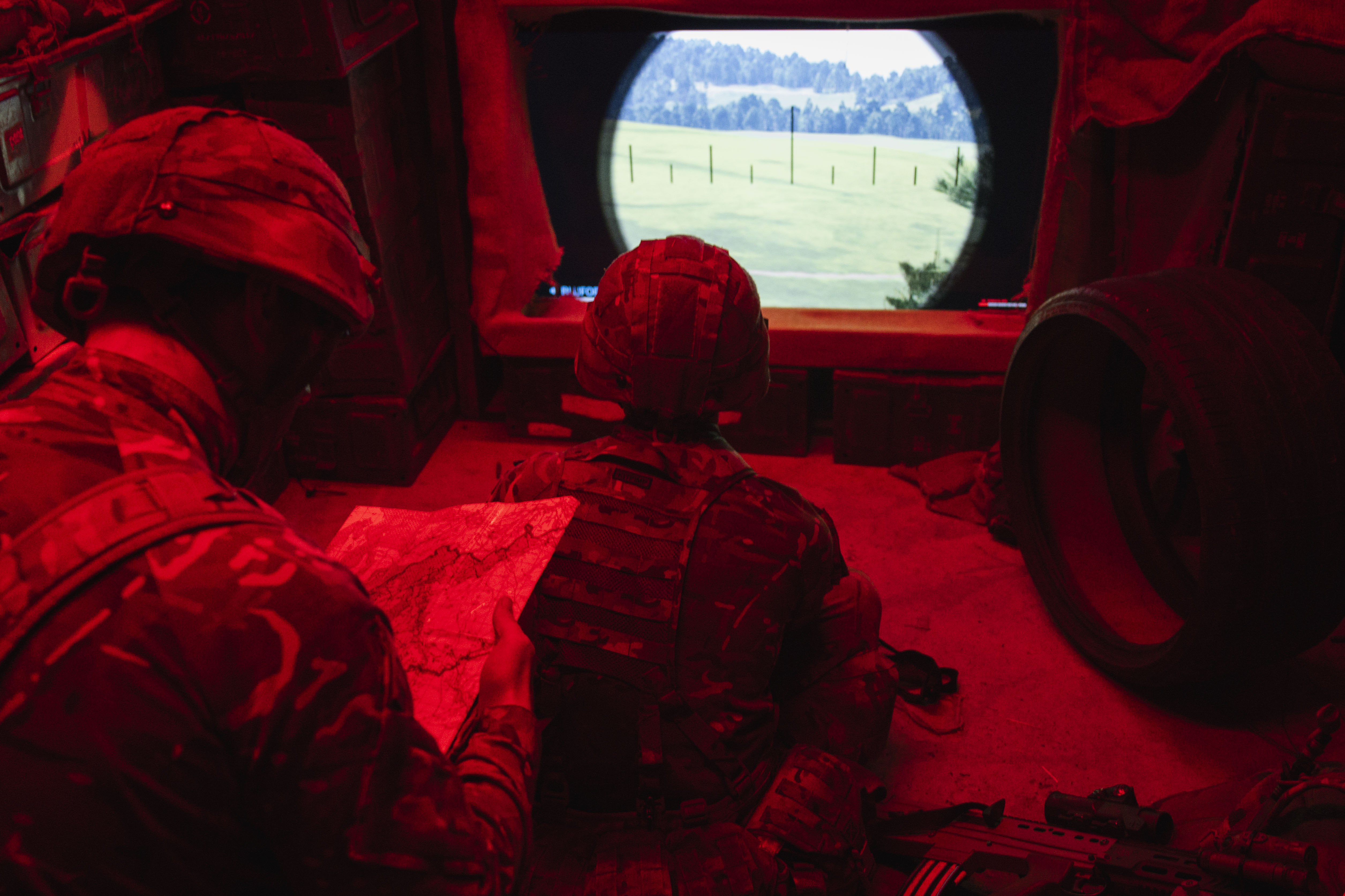 Two soldiers in a dimly lit room with red lighting study a map and a landscape through a screen