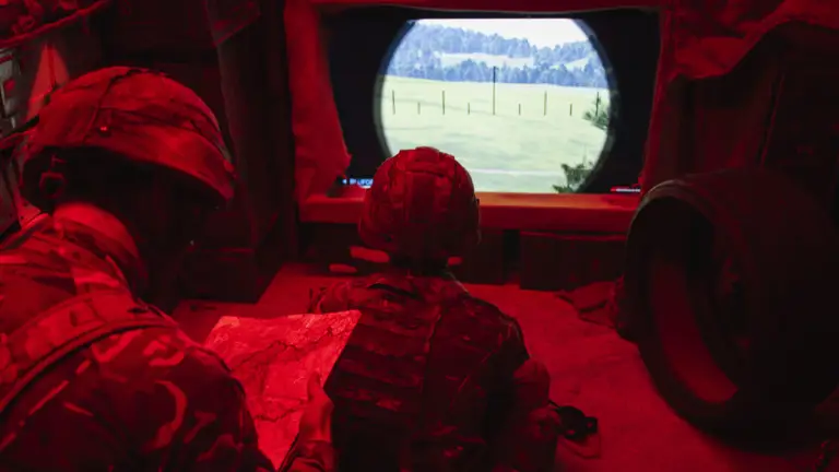 Two soldiers in a dimly lit room with red lighting study a map and a landscape through a screen