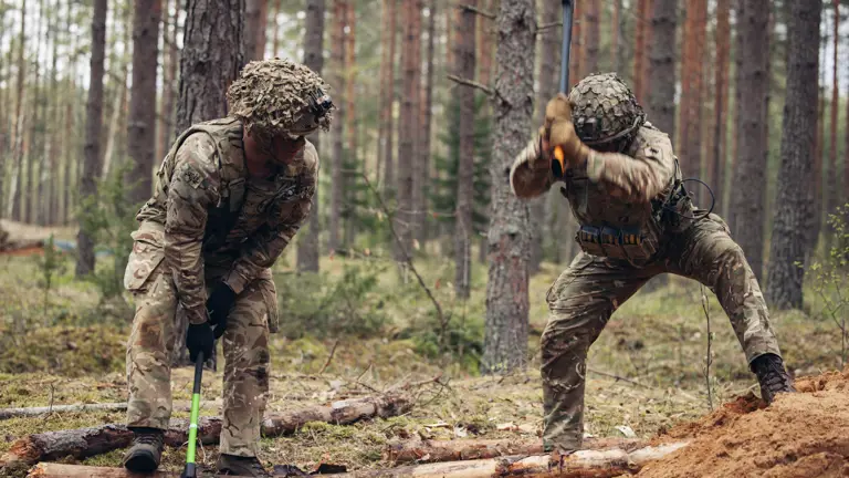 Two soldiers in uniform are pictured chopping wood with an axe.