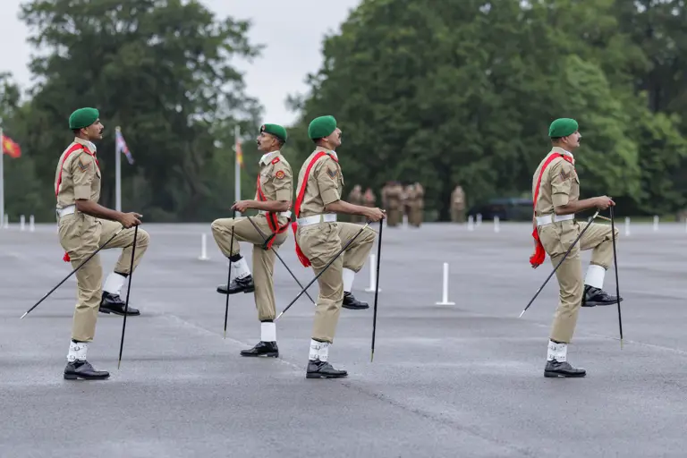 Marching individuals in uniform with green berets and red sashes are performing a synchronized drill on a large paved area. Trees are visible in the background, and a few other individuals are faintly seen in the distance.