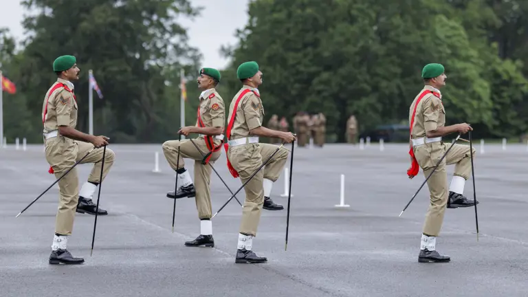 Marching individuals in uniform with green berets and red sashes are performing a synchronized drill on a large paved area. Trees are visible in the background, and a few other individuals are faintly seen in the distance.
