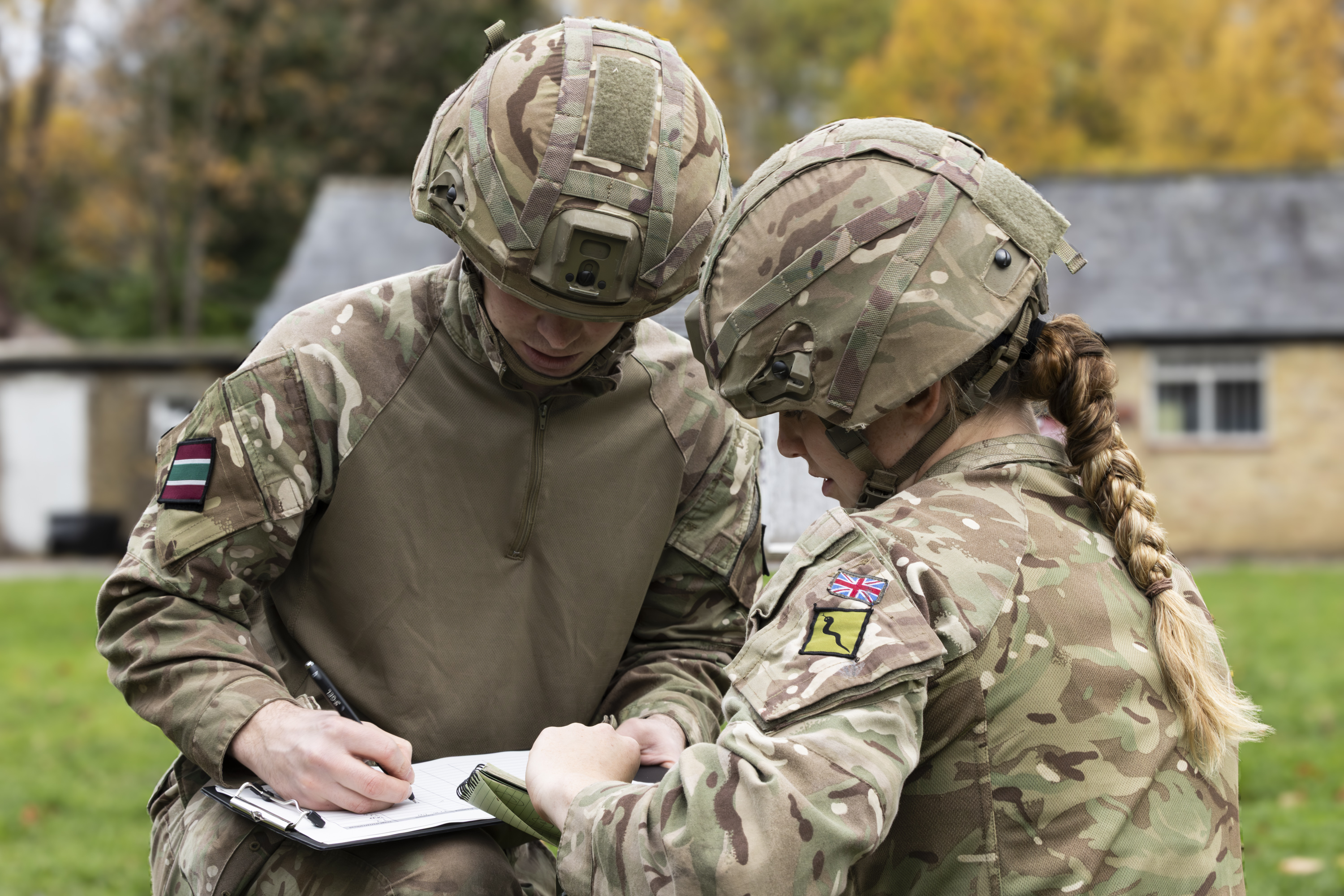 Two soldiers in camouflage uniforms and helmets review notes outdoors near a building with autumn trees.