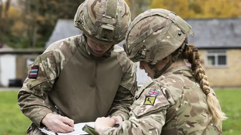 Two soldiers in camouflage uniforms and helmets review notes outdoors near a building with autumn trees.