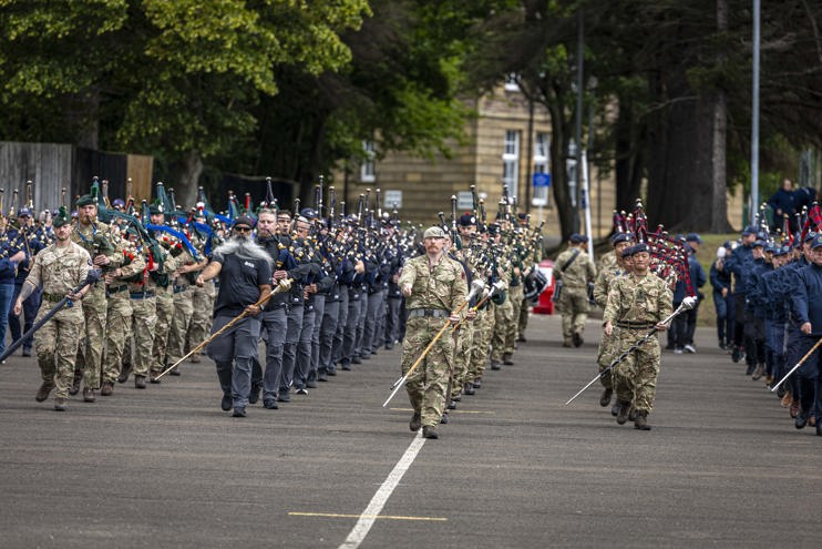 A group of individuals in military and ceremonial uniforms are marching down a parade square. They carry various instruments, including bagpipes and drums. Trees and buildings line the sides of the road, creating a structured backdrop for the procession.