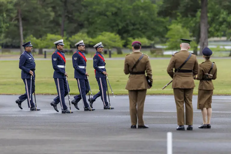 A group of four uniformed individuals in blue walk in formation across a paved area, holding swords. They are observed by three uniformed individuals in brown. The setting is an outdoor space with green grass and trees in the background.