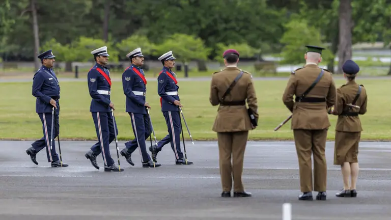 A group of four uniformed individuals in blue walk in formation across a paved area, holding swords. They are observed by three uniformed individuals in brown. The setting is an outdoor space with green grass and trees in the background.