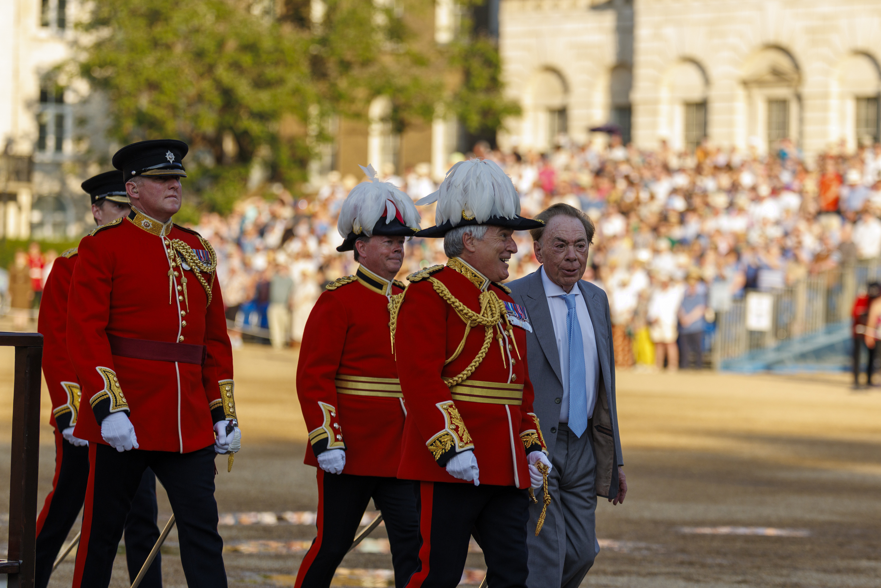 A group of people in formal red uniforms with gold detailing and white feathered hats sit together. One of them, possibly a public figure, is in focus. The background shows a blurred crowd, suggestive of a public event or ceremony.