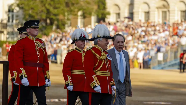 A group of people in formal red uniforms with gold detailing and white feathered hats sit together. One of them, possibly a public figure, is in focus. The background shows a blurred crowd, suggestive of a public event or ceremony.