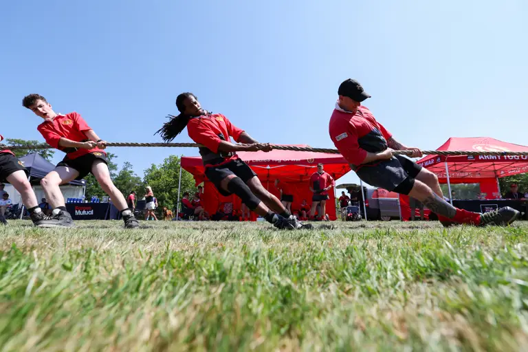 A group of individuals in red shirts participating in a tug-of-war game on a grassy field. Red Mutual tents are visible in the background under a clear blue sky.