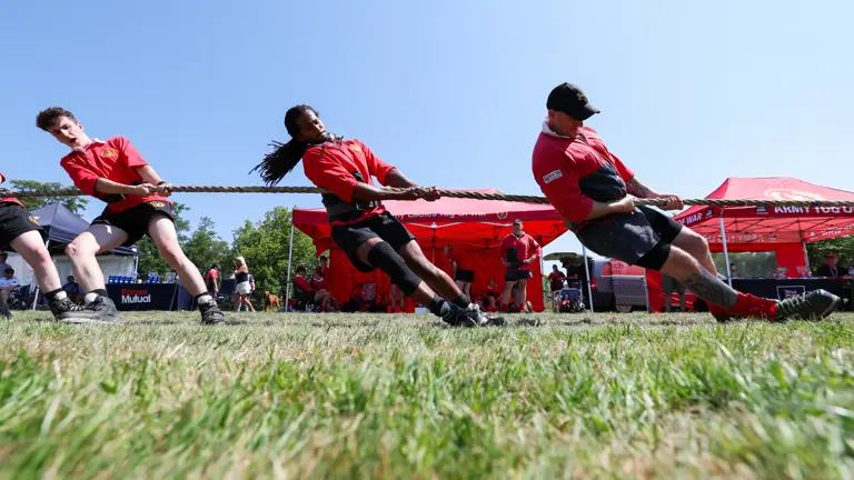 A group of individuals in red shirts participating in a tug-of-war game on a grassy field. Red Mutual tents are visible in the background under a clear blue sky.