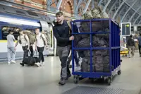 A man wearing a dark coat and grey hiking trousers pushes a blue trolley full of camouflage bags.