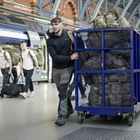 A man wearing a dark coat and grey hiking trousers pushes a blue trolley full of camouflage bags.