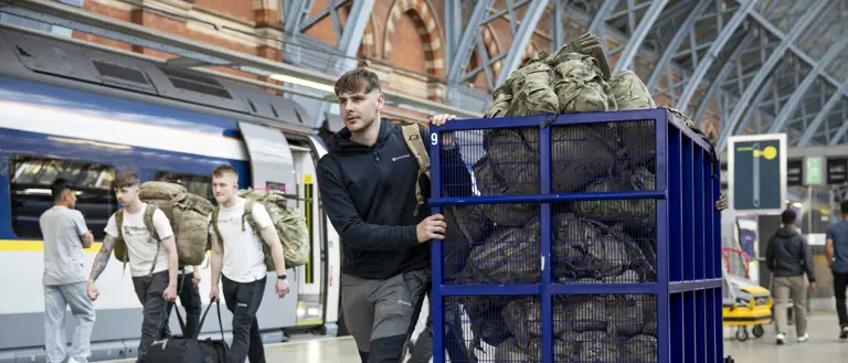 A man wearing a dark coat and grey hiking trousers pushes a blue trolley full of camouflage bags.