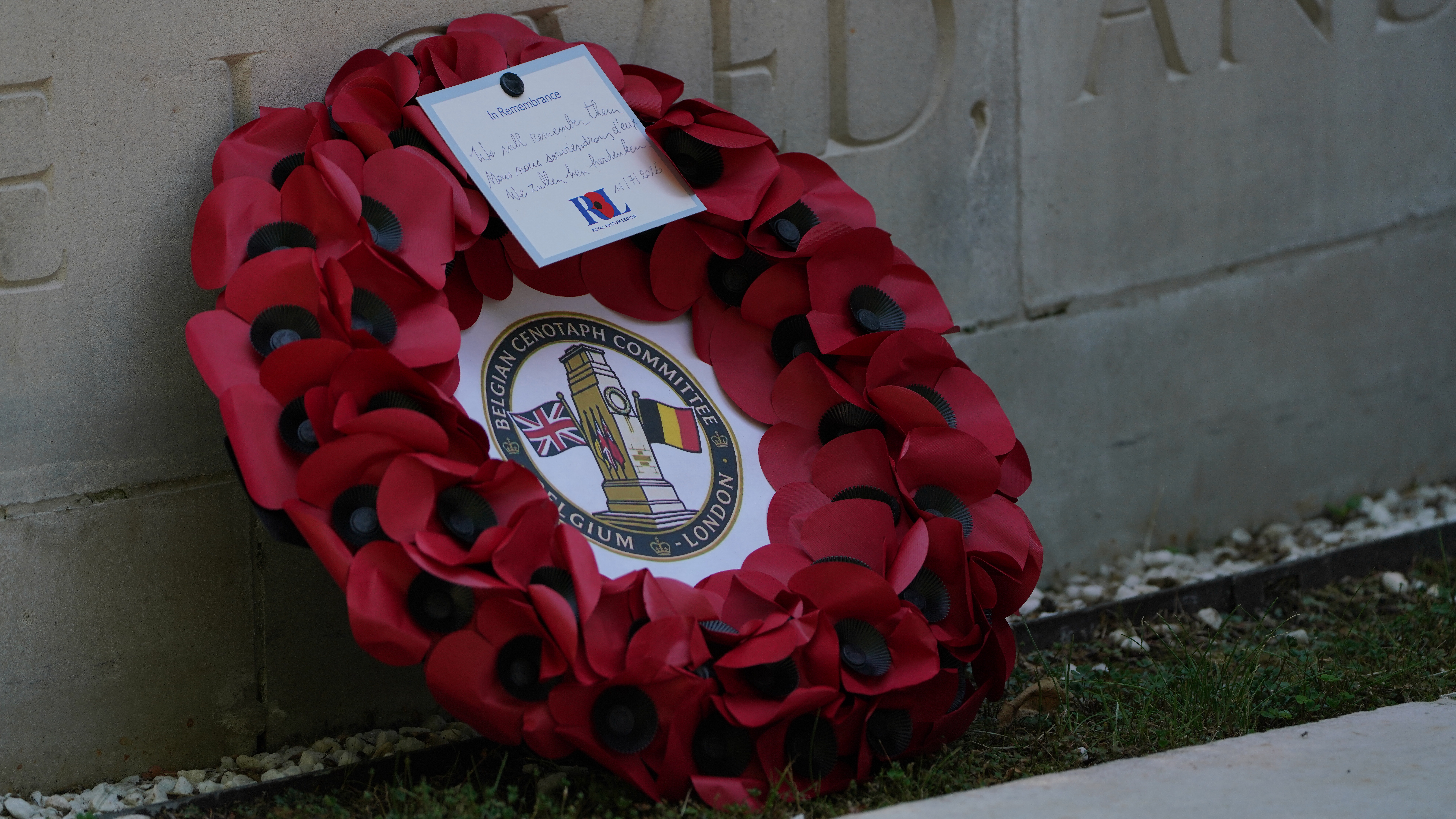 A wreath of red poppies is placed against a stone memorial. The wreath features a central emblem with two flags.