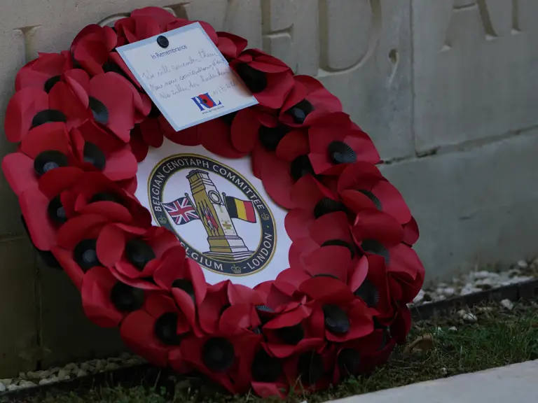 A wreath of red poppies is placed against a stone memorial. The wreath features a central emblem with two flags.