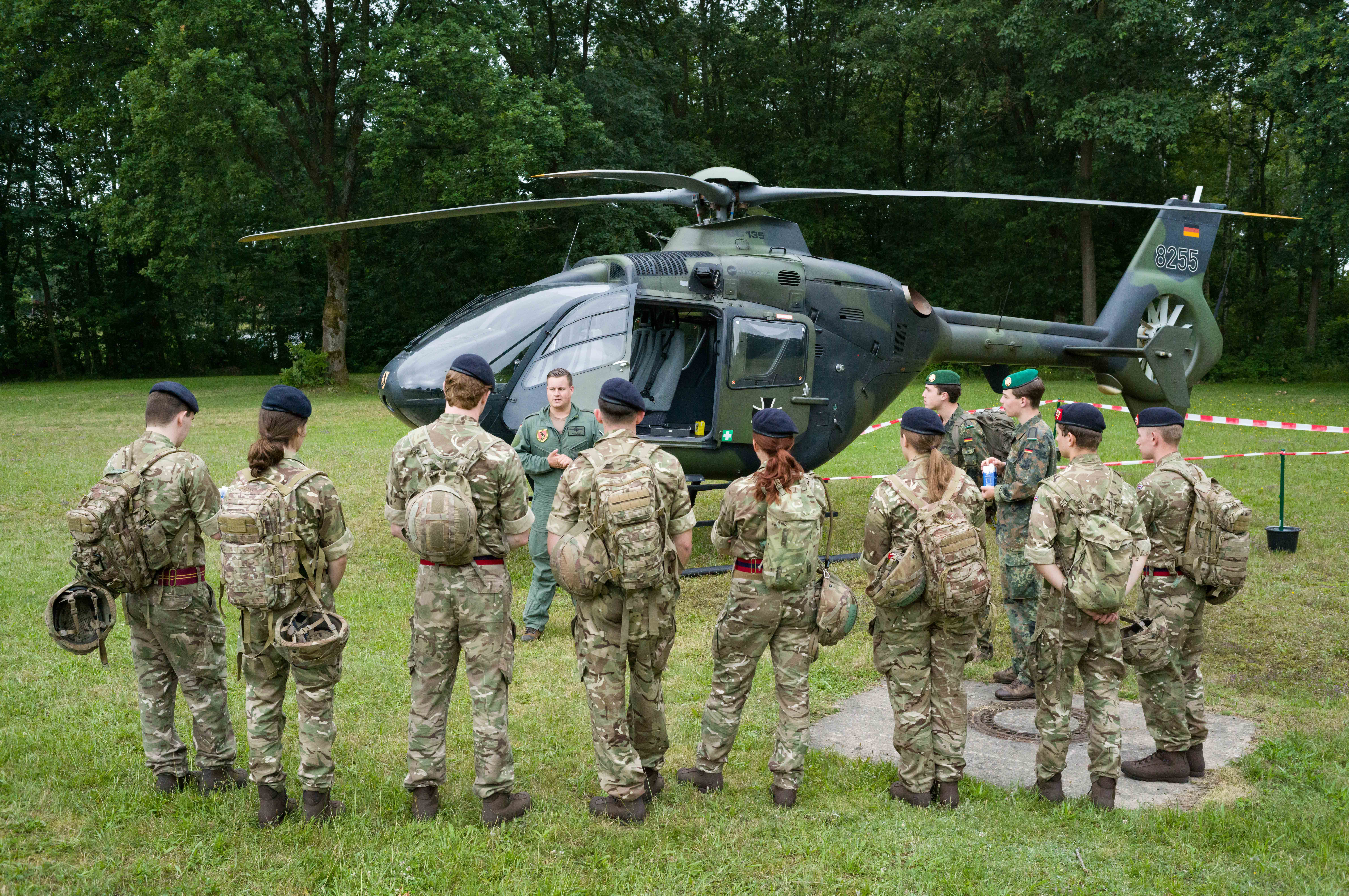 A group of people in military uniforms stand in a semi-circle on a grassy field, facing an instructor. Behind them is a military helicopter parked near a wooded area.