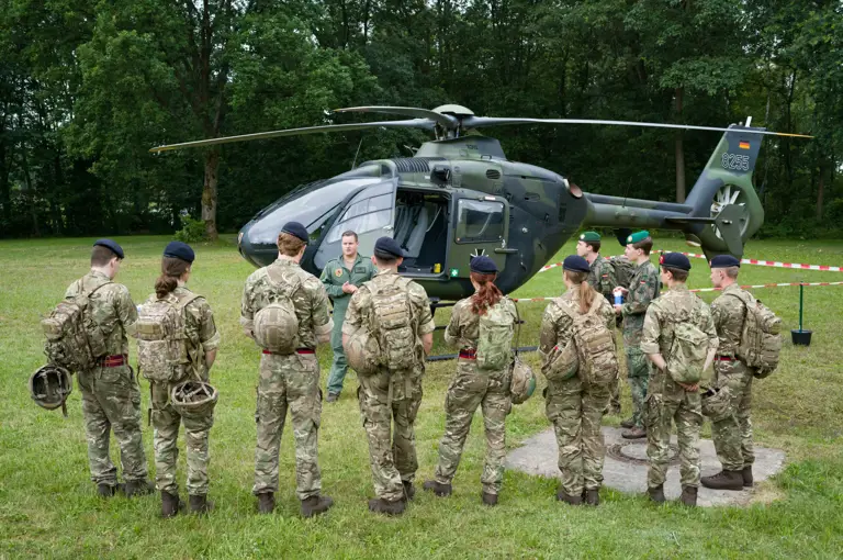 A group of people in military uniforms stand in a semi-circle on a grassy field, facing an instructor. Behind them is a military helicopter parked near a wooded area.