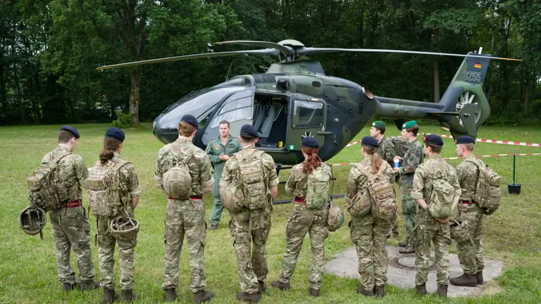 A group of people in military uniforms stand in a semi-circle on a grassy field, facing an instructor. Behind them is a military helicopter parked near a wooded area.