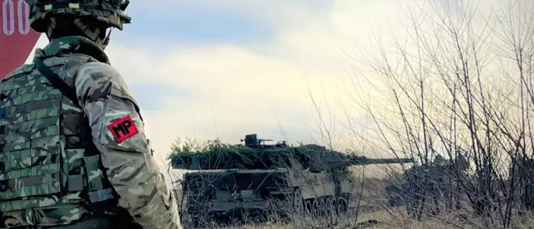 Military police soldier in camouflage gear stands guard near a red sign with armored vehicles in a grassy field.