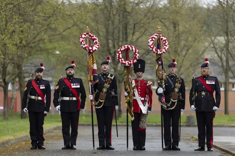 Soldiers in black and red tunics hold banners while stood on parade.