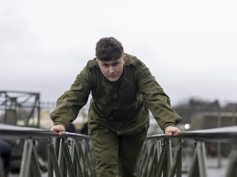 Person in green military uniform carefully walking across a narrow metal bridge outdoors on a cloudy day.