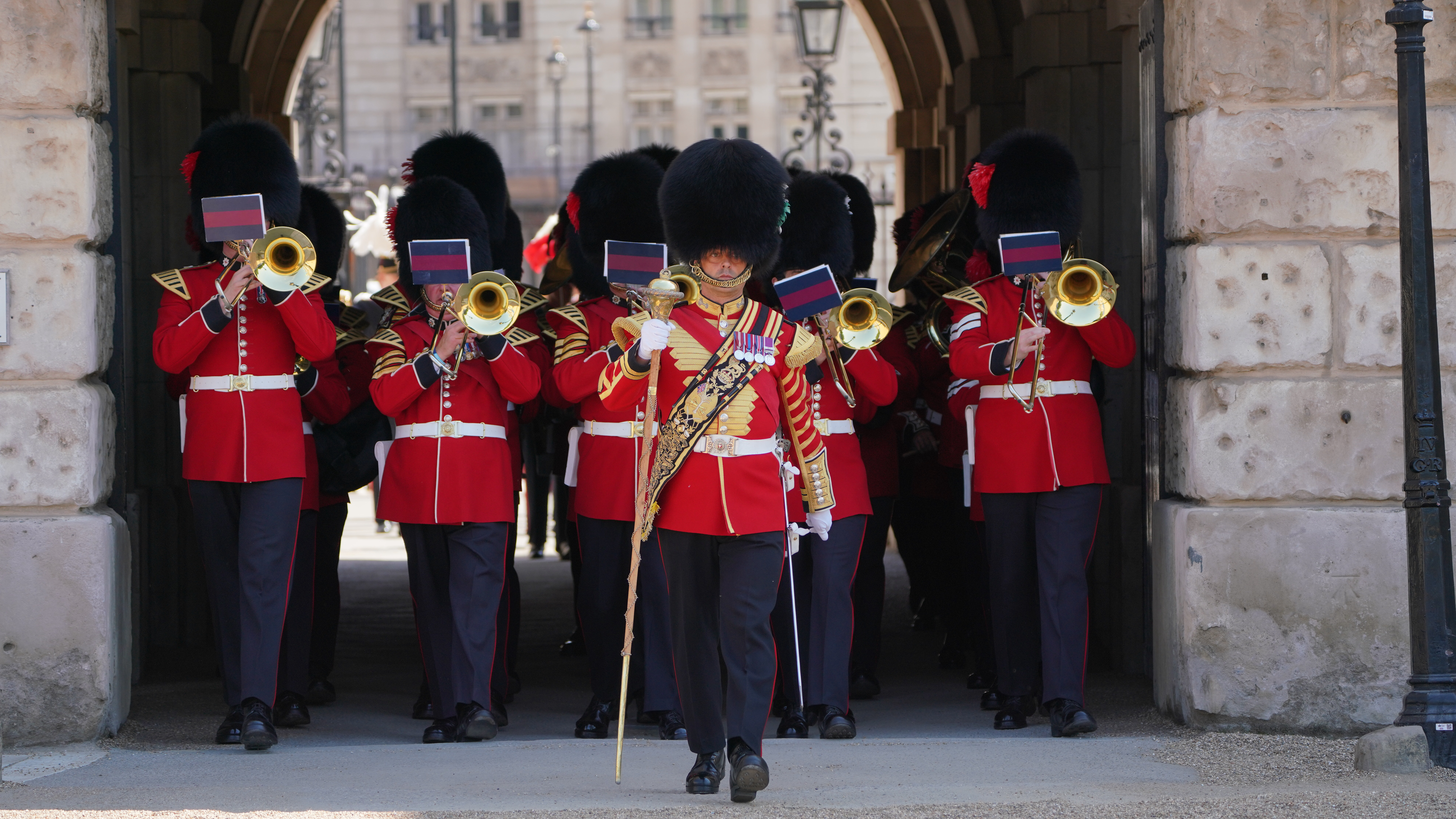 A group of marching band members wearing red uniforms and tall black hats plays brass instruments. 