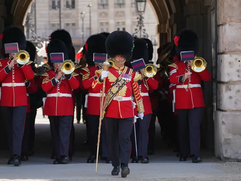 A group of marching band members wearing red uniforms and tall black hats plays brass instruments.