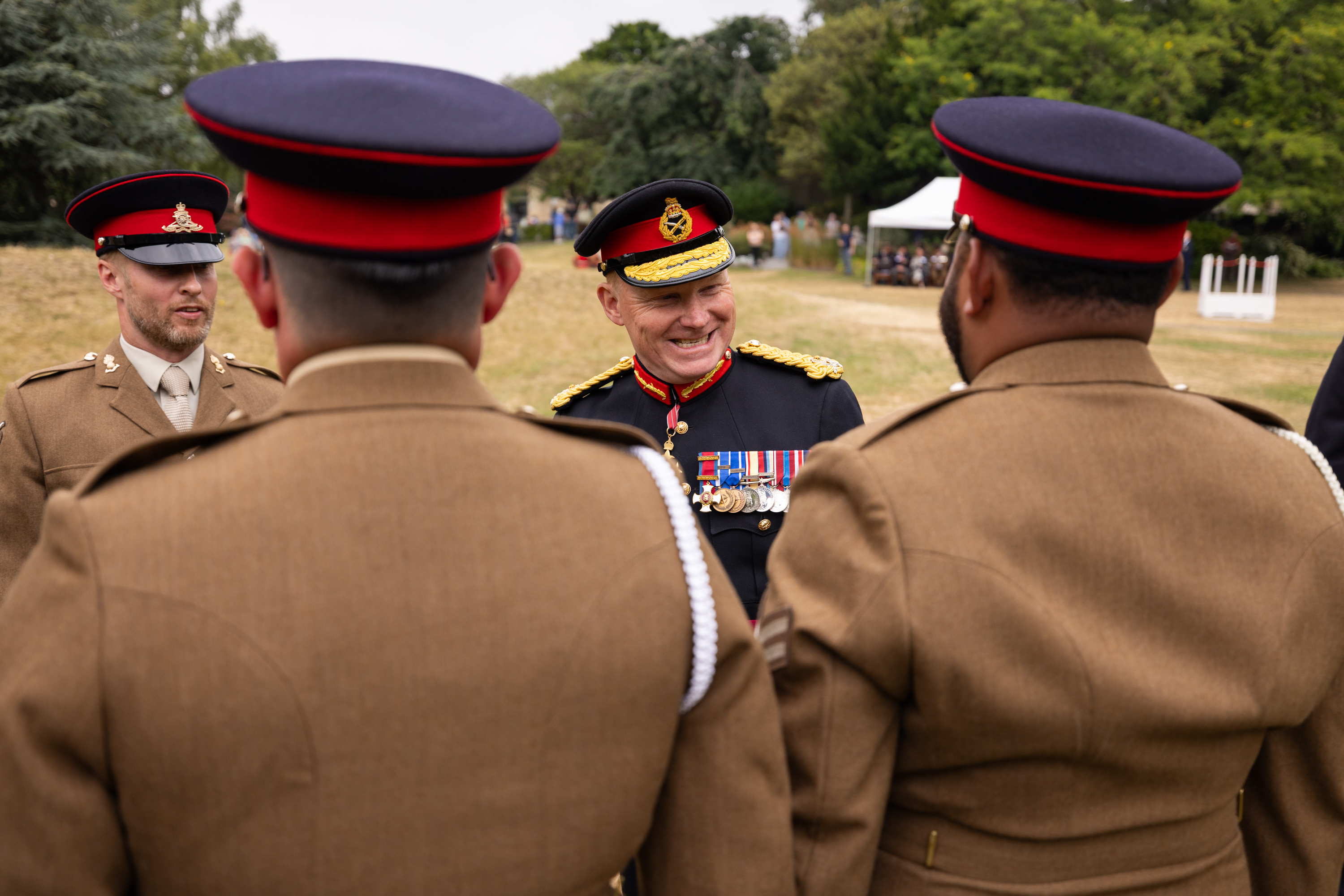 A group of individuals in military uniforms, with one individual facing the others and smiling. The scene is outdoors.