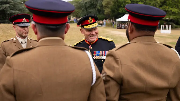 A group of individuals in military uniforms, with one individual facing the others and smiling. The scene is outdoors.