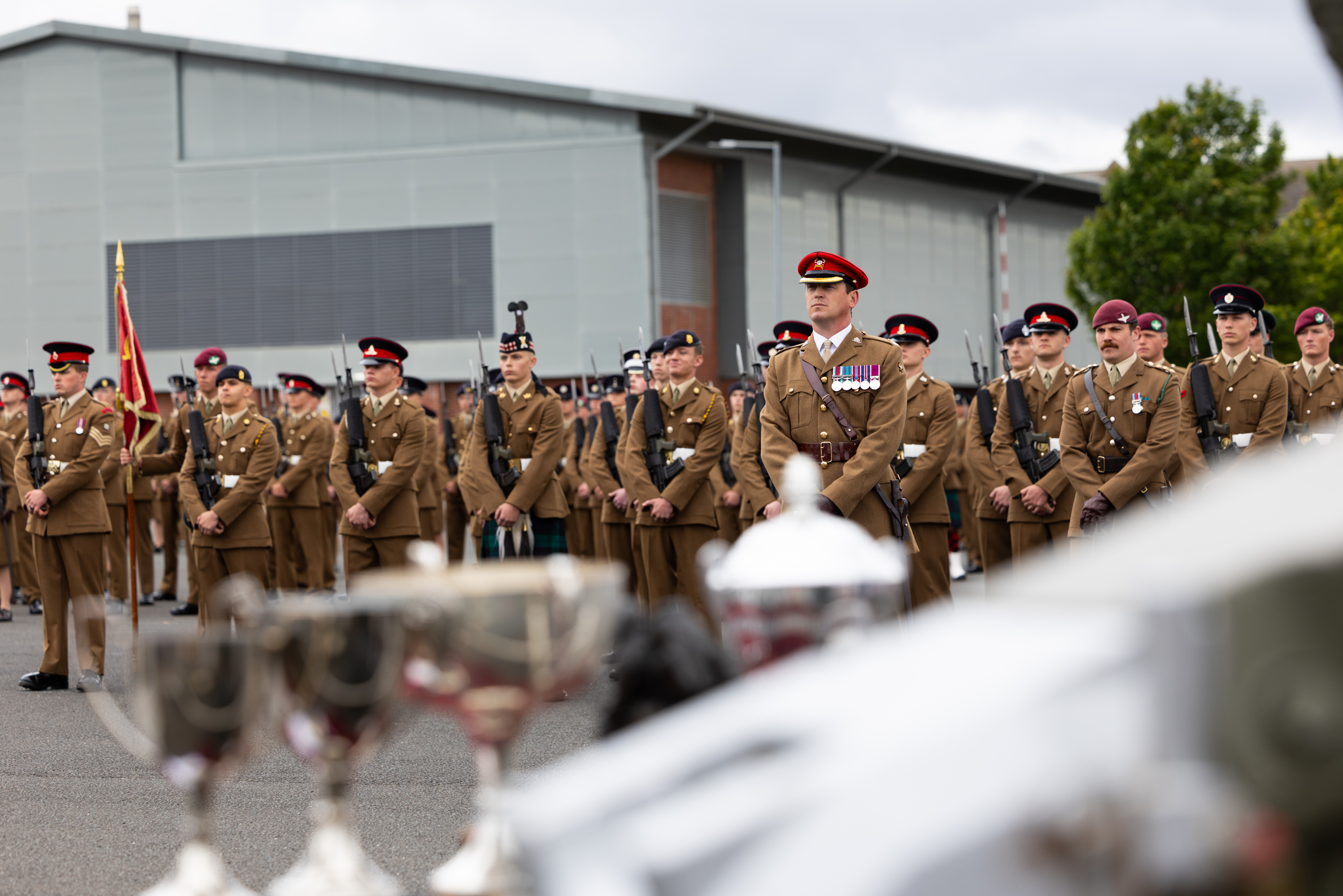 A group of uniformed military personnel stand in formation during a ceremony. Some are holding a flag. Several members have medals on their uniforms. In the foreground, blurred trophies are visible. A modern building is in the background.