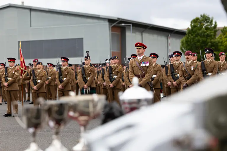 A group of uniformed military personnel stand in formation during a ceremony. Some are holding a flag. Several members have medals on their uniforms. In the foreground, blurred trophies are visible. A modern building is in the background.