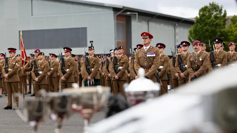 A group of uniformed military personnel stand in formation during a ceremony. Some are holding a flag. Several members have medals on their uniforms. In the foreground, blurred trophies are visible. A modern building is in the background.