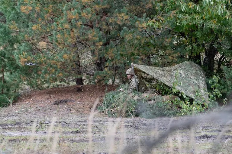 A soldier kneels under a camouflage shelter.