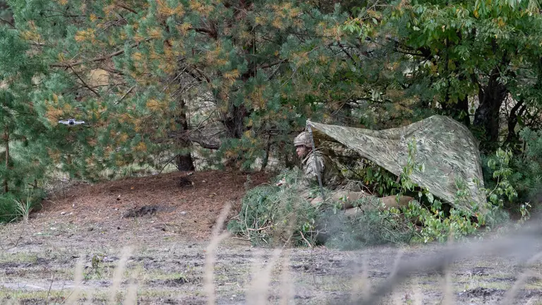 A soldier kneels under a camouflage shelter.
